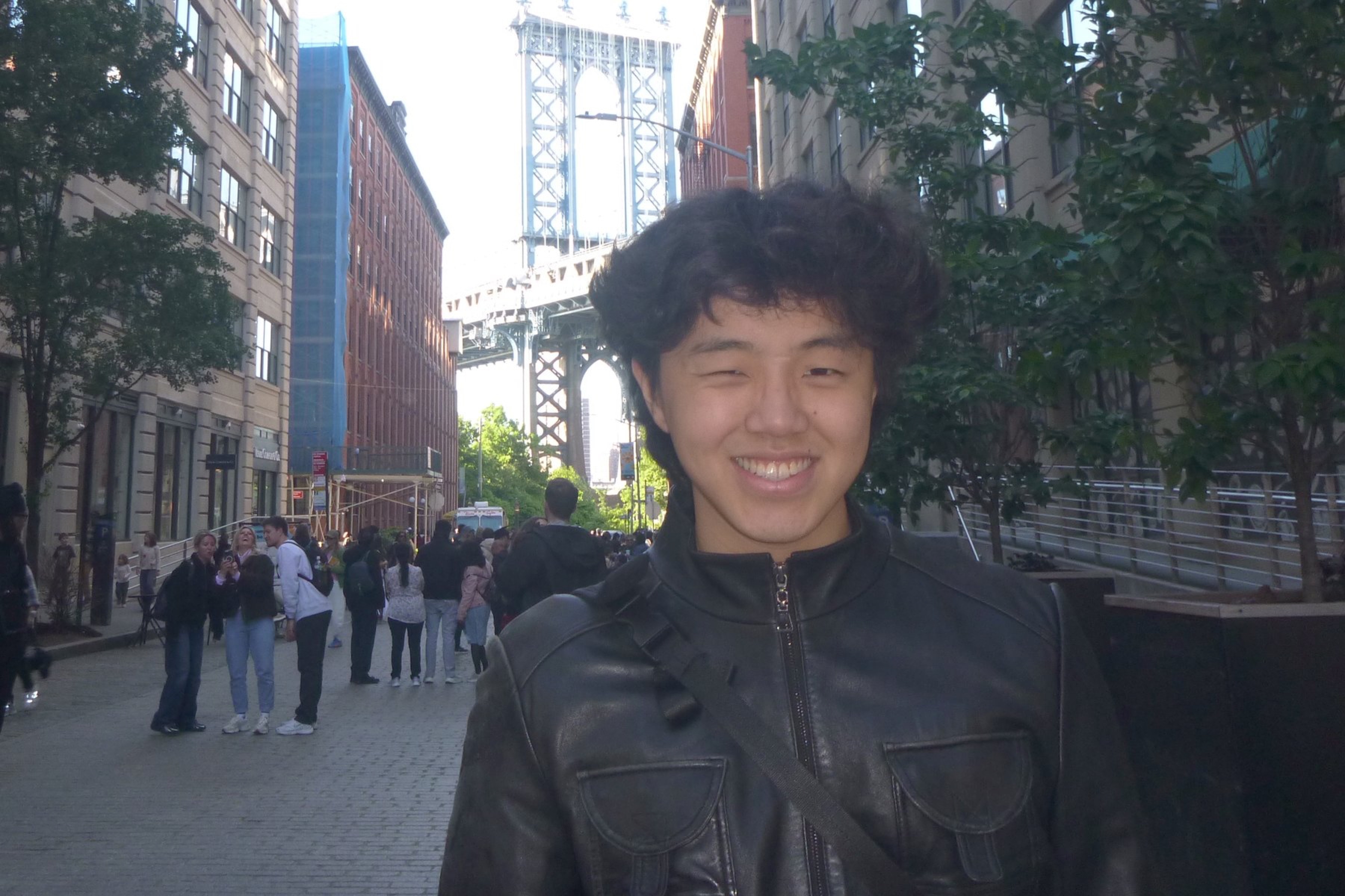 Daniel smiling on a cobbled street in Dumbo, in a black leather jacket, the Manhattan Bridge rising between the buildings directly behind him