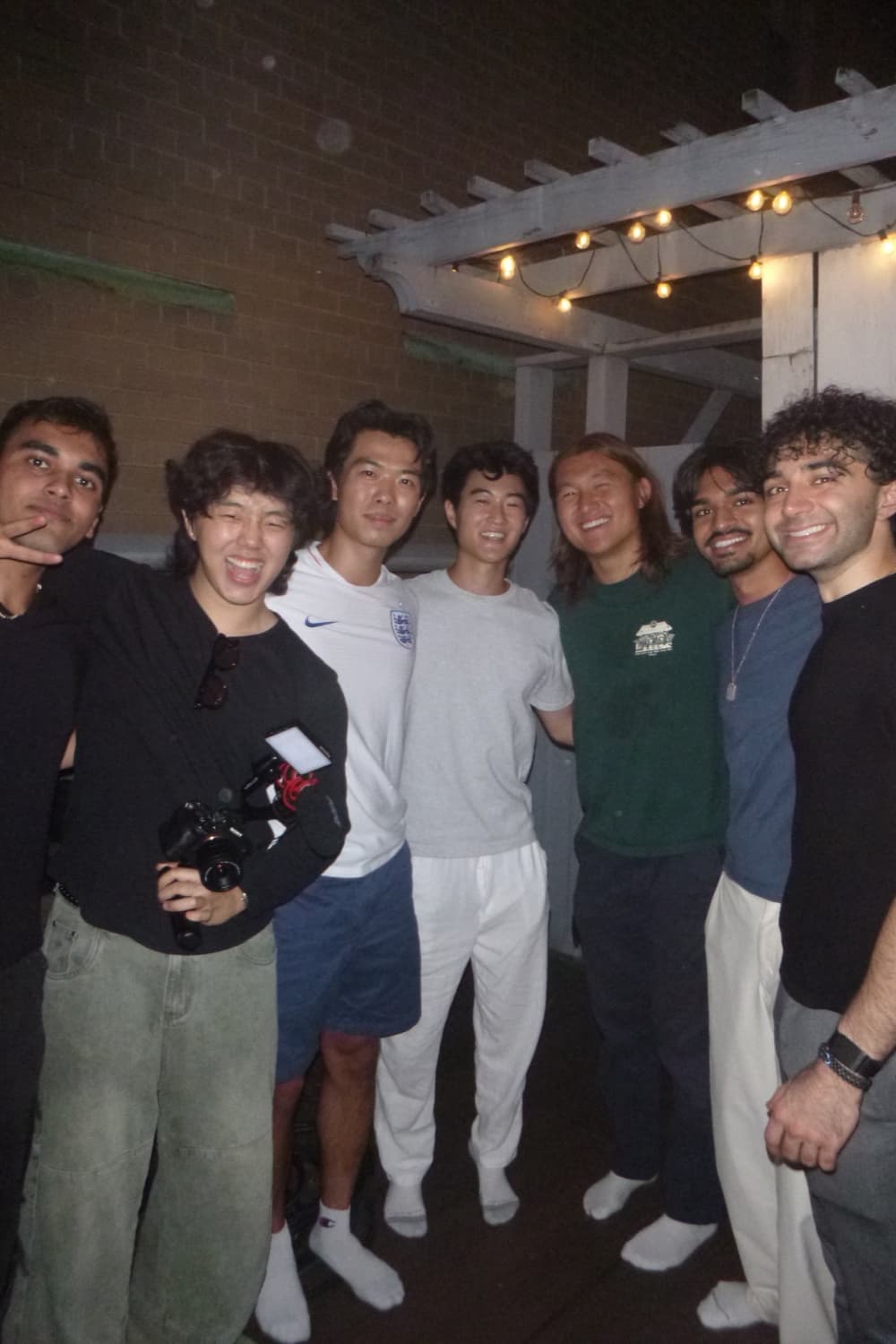 Eight friends crowded together on a balcony at night under string lights, Daniel second from left holding a camera