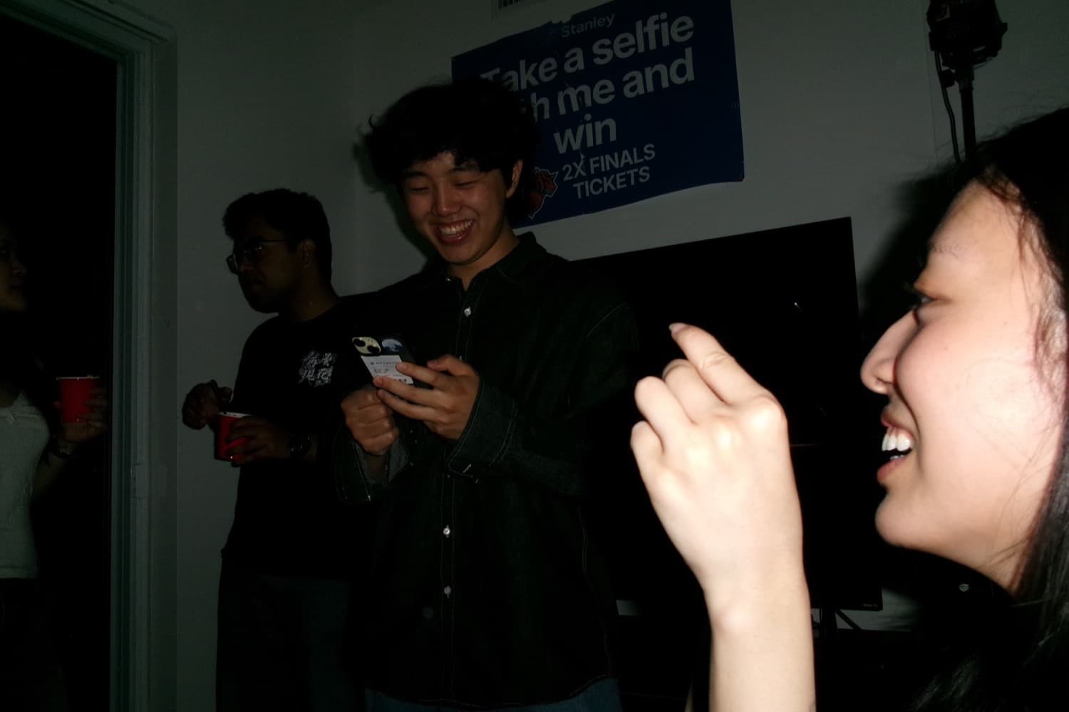 Daniel laughing at his phone at a crowded indoor event, in front of a poster reading take a selfie with me and win 2X FINALS TICKETS