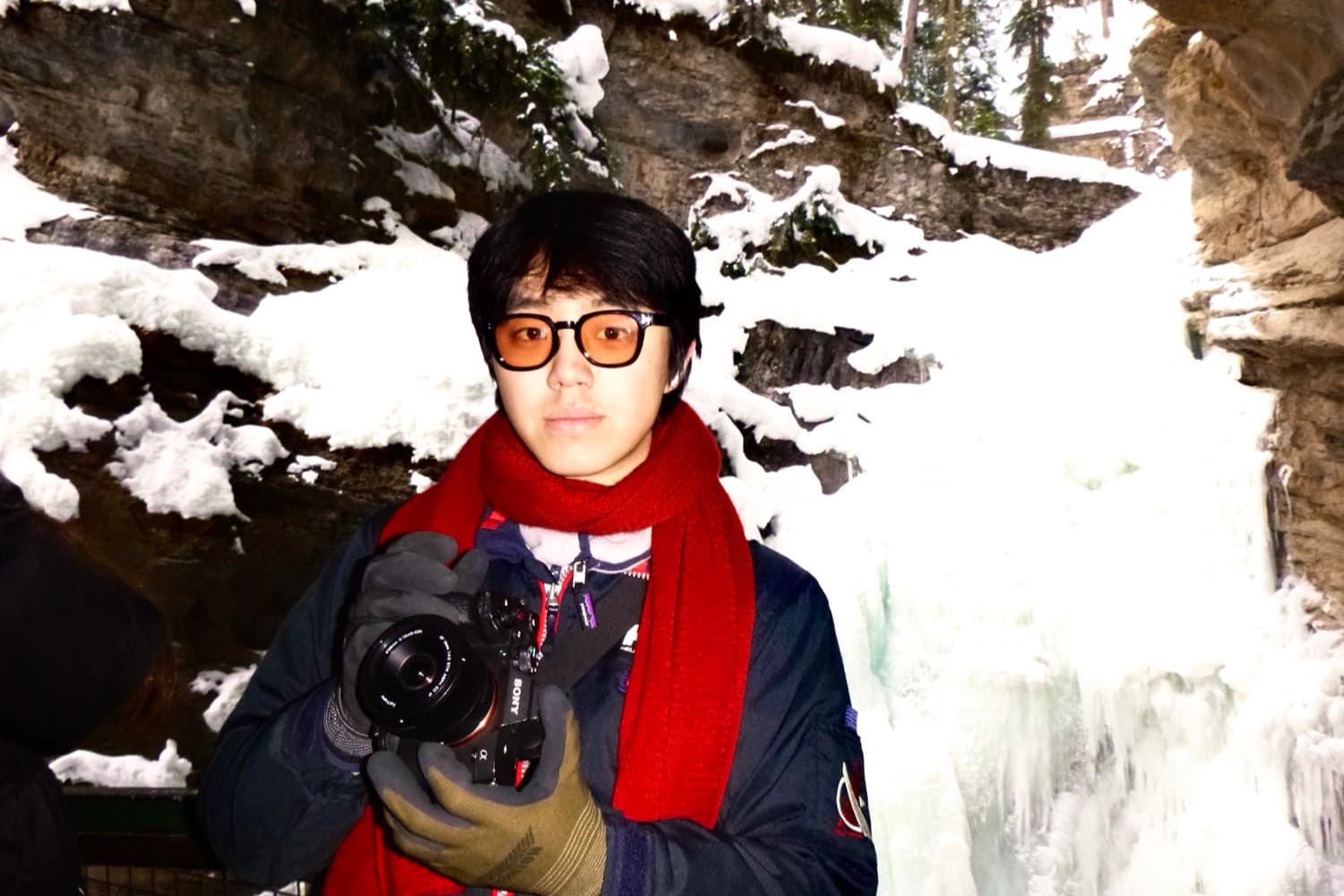Daniel in a parka and red scarf holding a camera, standing in a canyon of frozen rock and ice