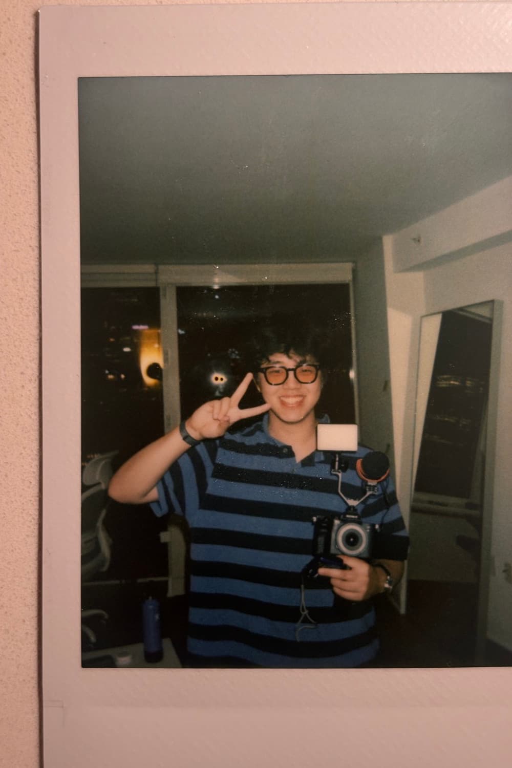 An instax print of Daniel in a striped polo and round glasses, holding a film camera and making a peace sign