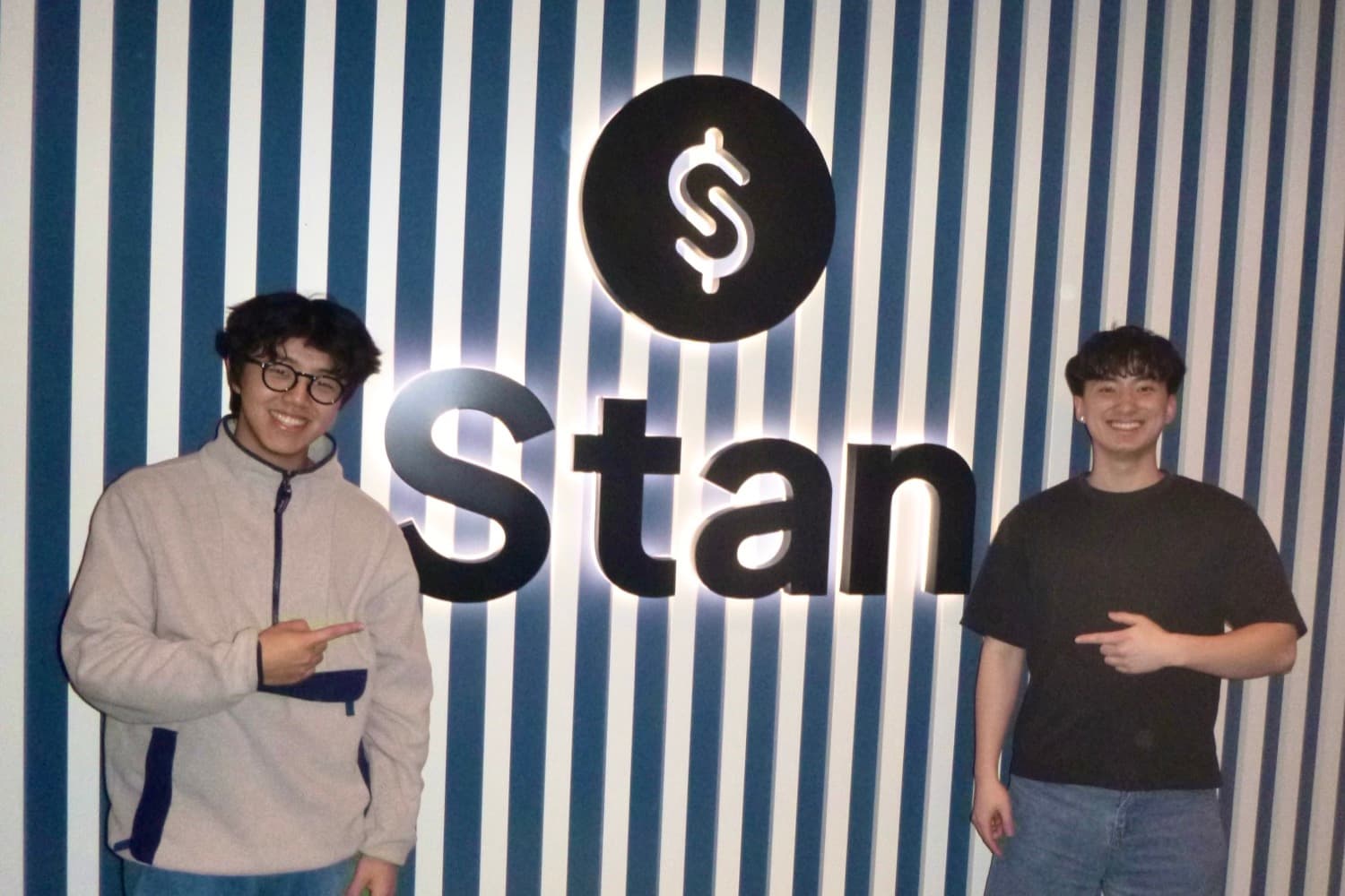 Daniel and a colleague standing either side of an illuminated Stan sign on a blue and white striped wall, both pointing at it